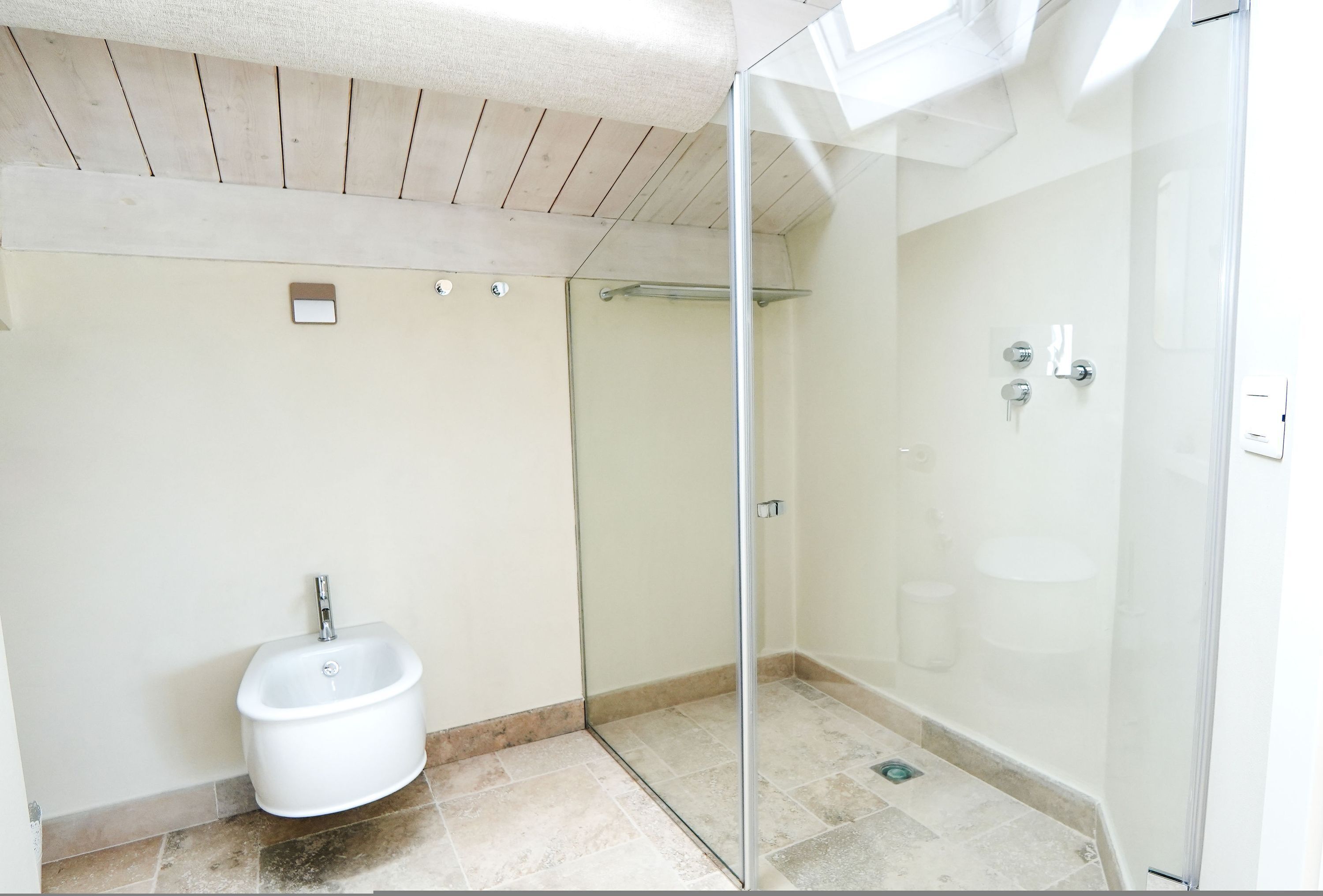 A modern bathroom with a glass shower enclosure, a white pedestal bidet, and a tiled floor.