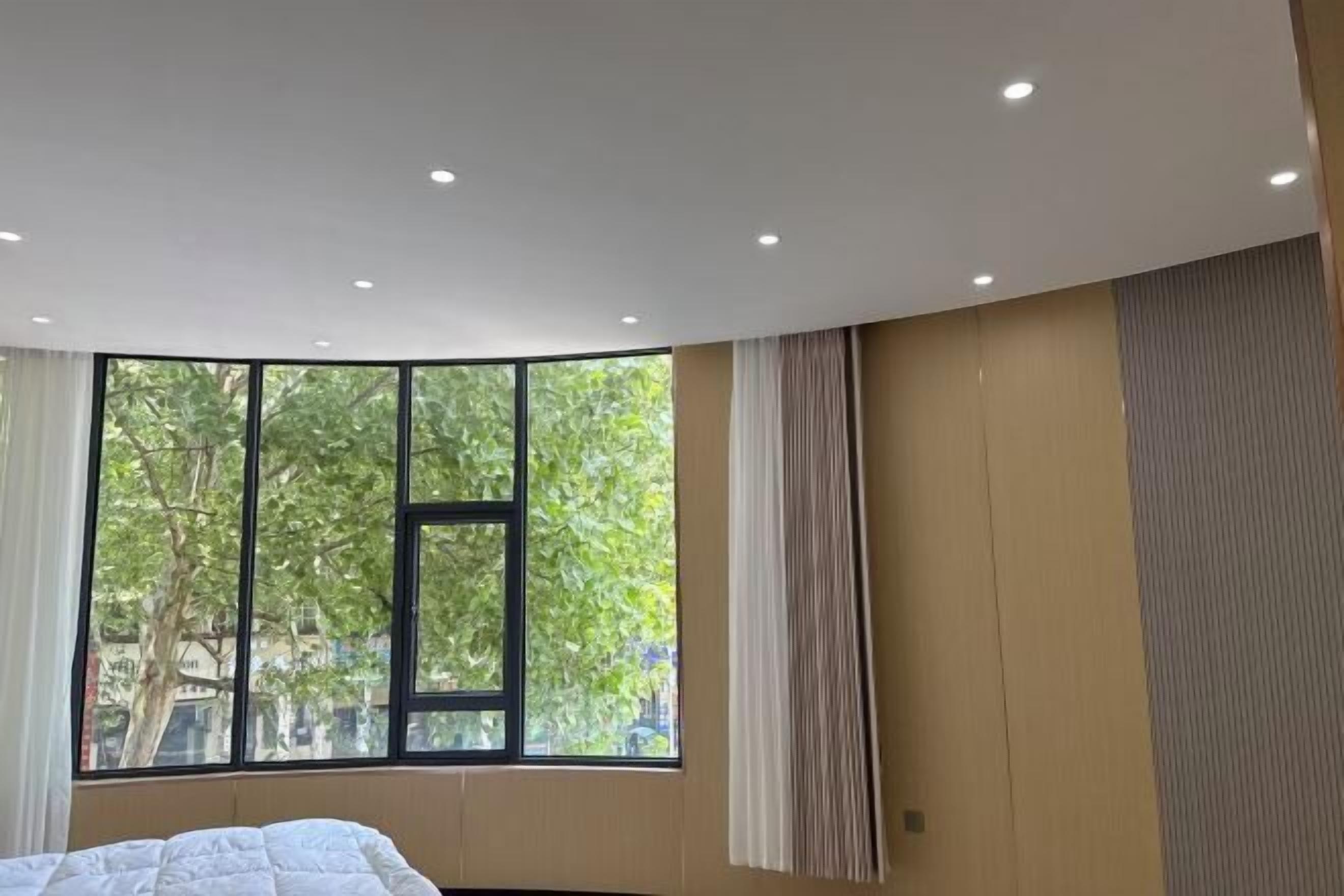A modern hotel room with a large window, a bed, and a ceiling with recessed lighting.