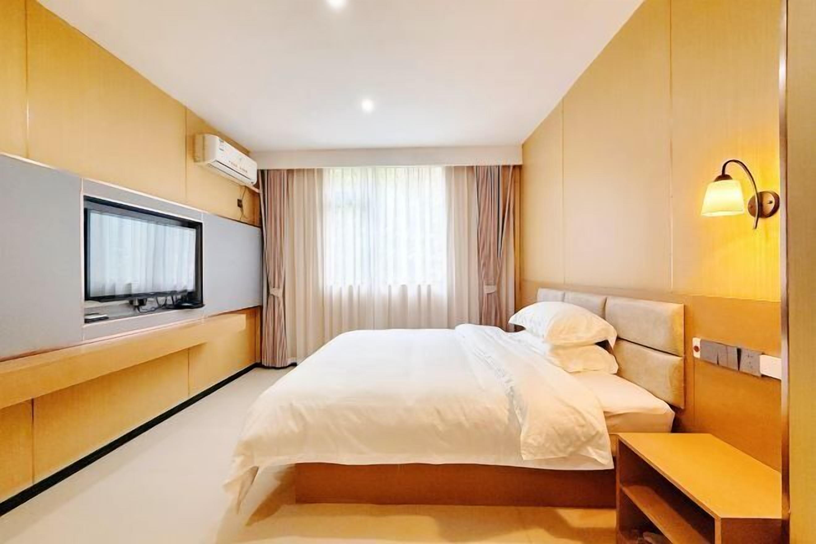 A modern hotel room with a large bed, a flat-screen TV, a bedside table, and a wall-mounted lamp.