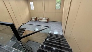 Ein modernes Interieur mit einer Wendeltreppe aus Glasgeländer, einer Couch und zwei Sesseln in einem Zimmer mit großen Fenstern und Fliesenboden.