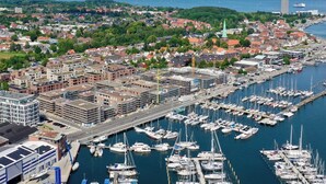 En småbåtshamn med ett stort antal förtöjda båtar, omgiven av en blandning av bostads- och affärsbyggnader, inklusive ett framträdande kyrktorn.