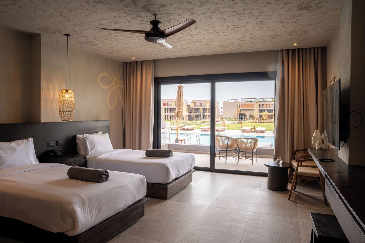 A hotel room with two beds, a ceiling fan, a TV, and a view of a swimming pool and buildings.