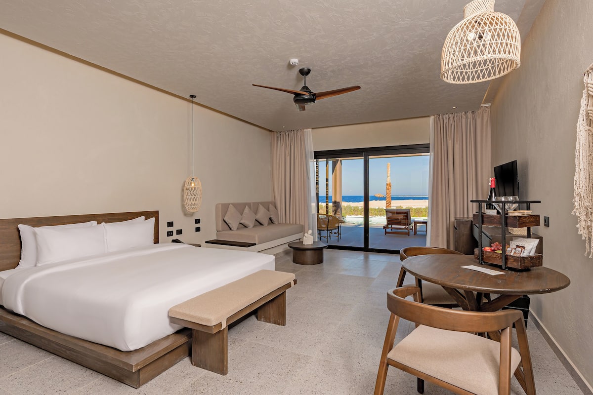 A hotel room with a large bed, a ceiling fan, a dining table with chairs, and a view of the ocean through the window.