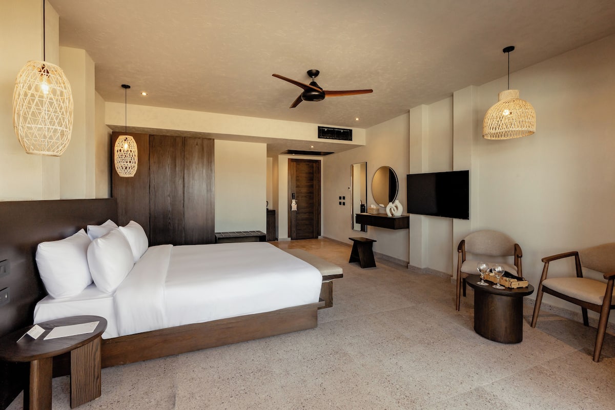 A modern hotel room with a large bed, a ceiling fan, a TV, and two chairs.