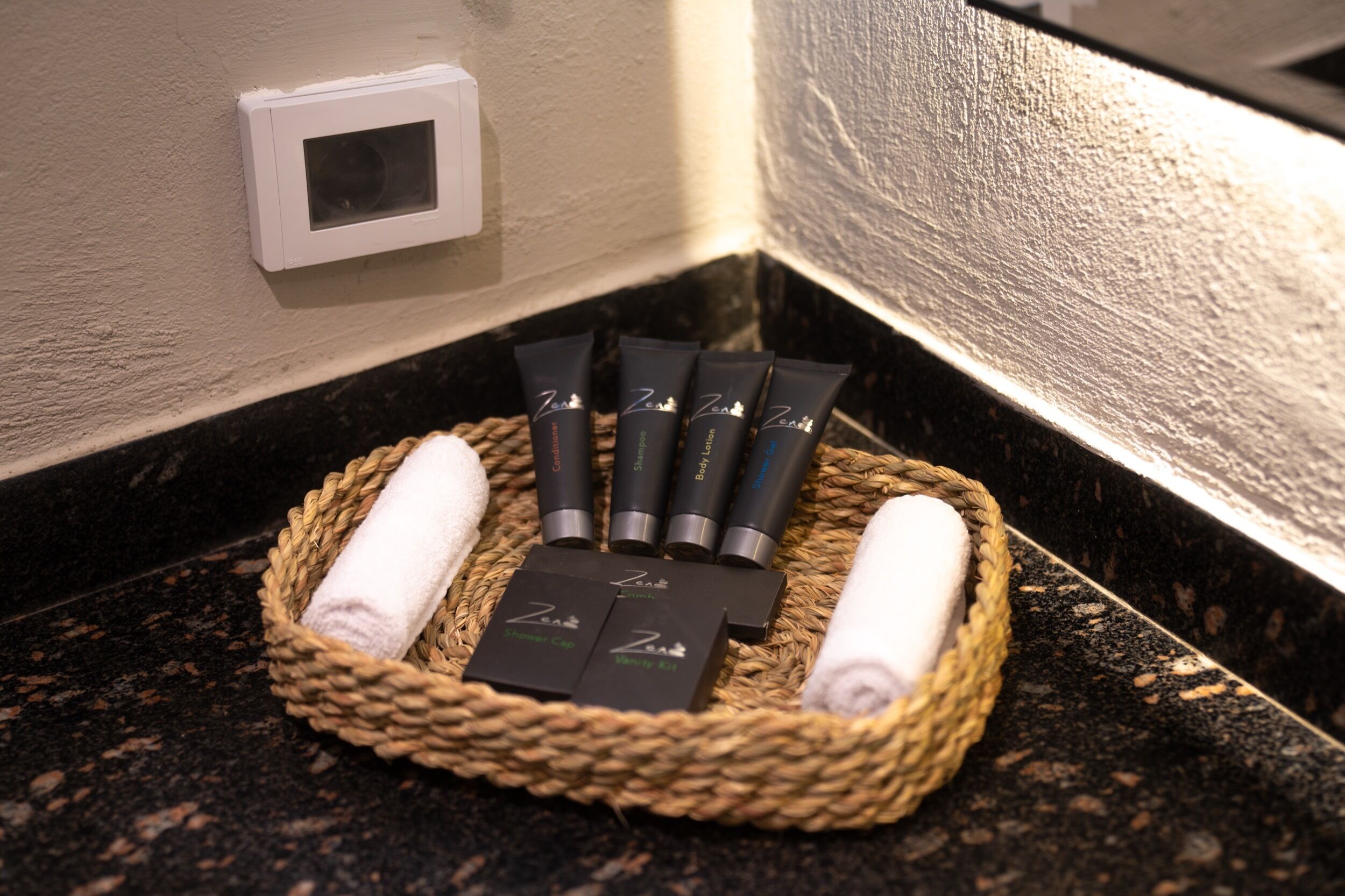 A bathroom corner with a woven basket containing toiletries, a towel, and a small white box.