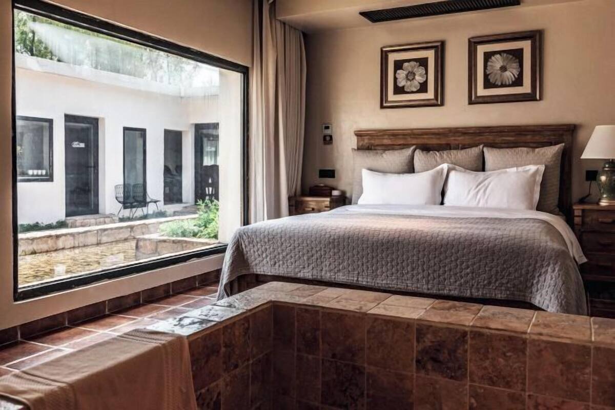 A hotel room with a large window, a bed with a wooden headboard, and two framed floral pictures on the wall.