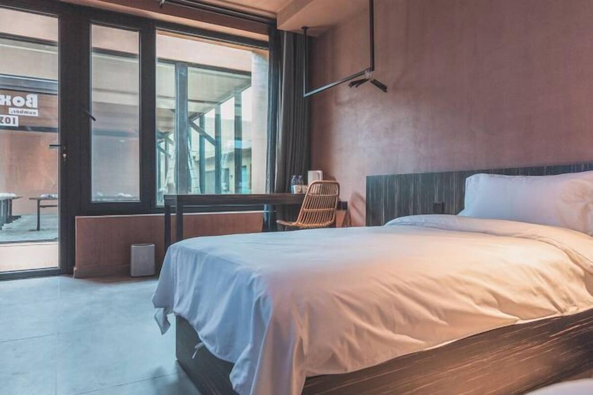 A modern hotel room with a large bed, a chair, a table, and a view through a glass door.