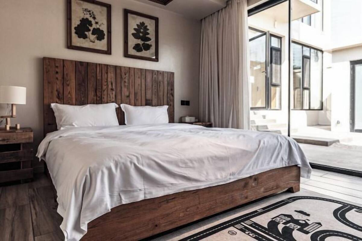 A bedroom with a wooden headboard, white bedding, a nightstand with a lamp, and a large window with curtains.