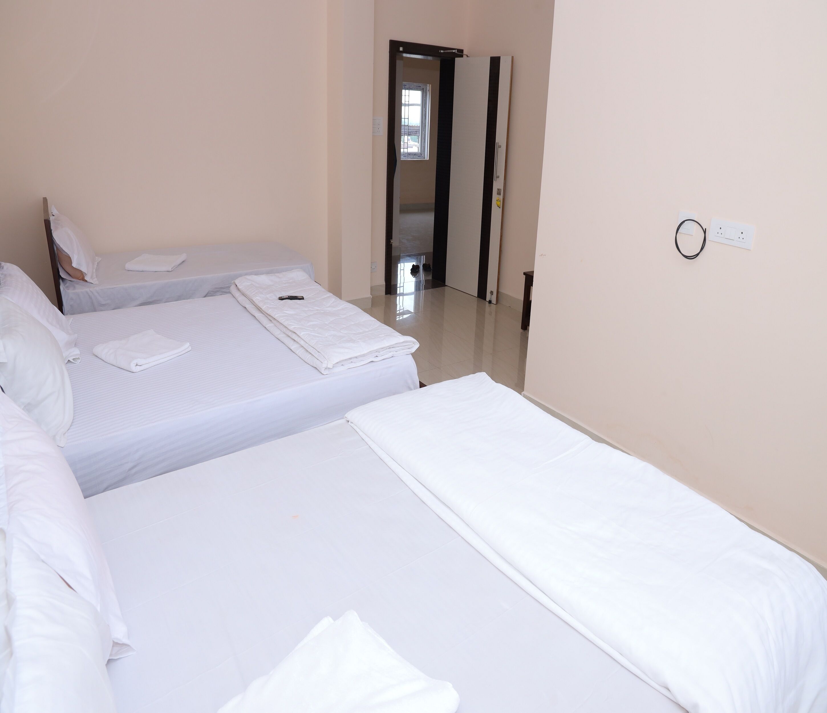 A hotel room with three beds arranged in a row, a door leading to a hallway, and a power outlet on the wall.