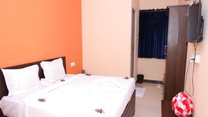 Una cama bien hecha con sábanas blancas y flores decorativas, una mesita de noche de madera con un arreglo floral rojo y blanco, y un televisor montado en la pared.