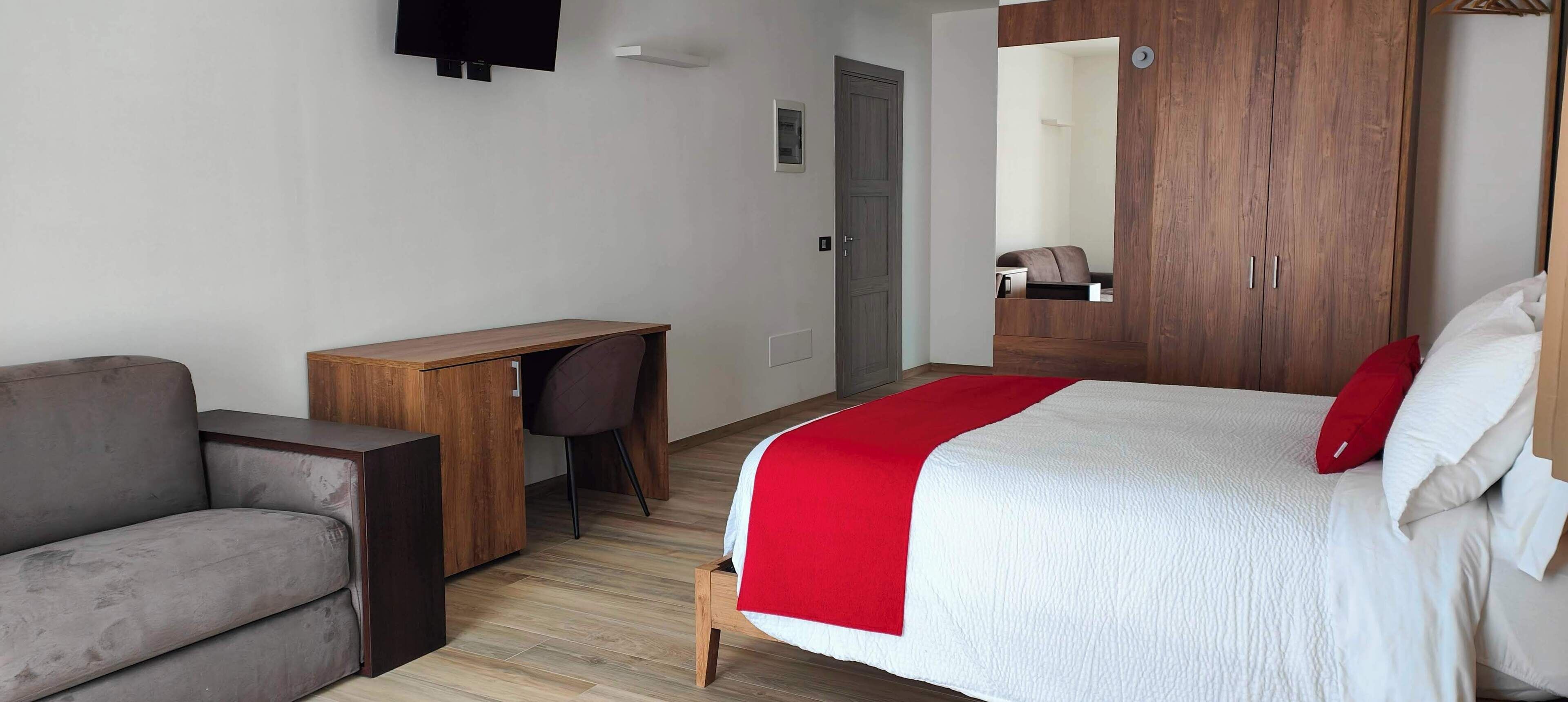 A hotel room with a bed, a desk, a chair, a sofa, a wardrobe, and a television.