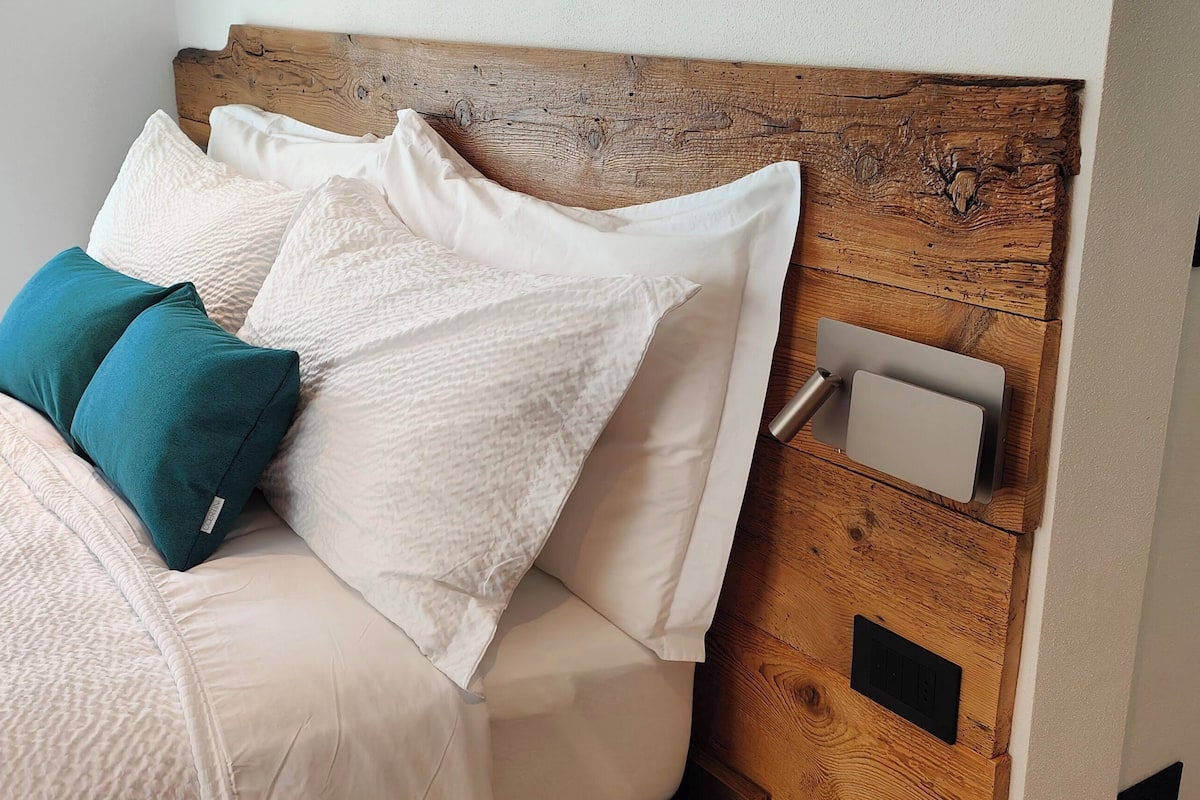 Een bed met een houten hoofdbord, witte en turquoise kussens en een nachtkastje met een lichtschakelaar.
