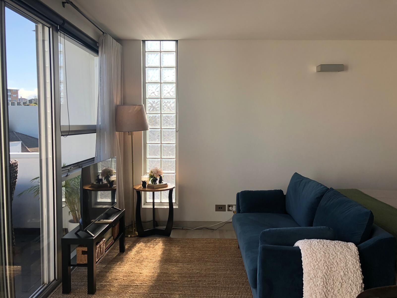 Een woonkamer met een blauwe bank, een klein bijzettafeltje met een lamp en een groot raam met uitzicht op gebouwen.