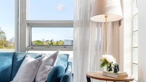 A room with a blue sofa, a round wooden side table with a lamp and flowers, and a view of the outdoors through a window with sheer curtains. - La Casa De Frida  (Bondi Beach)