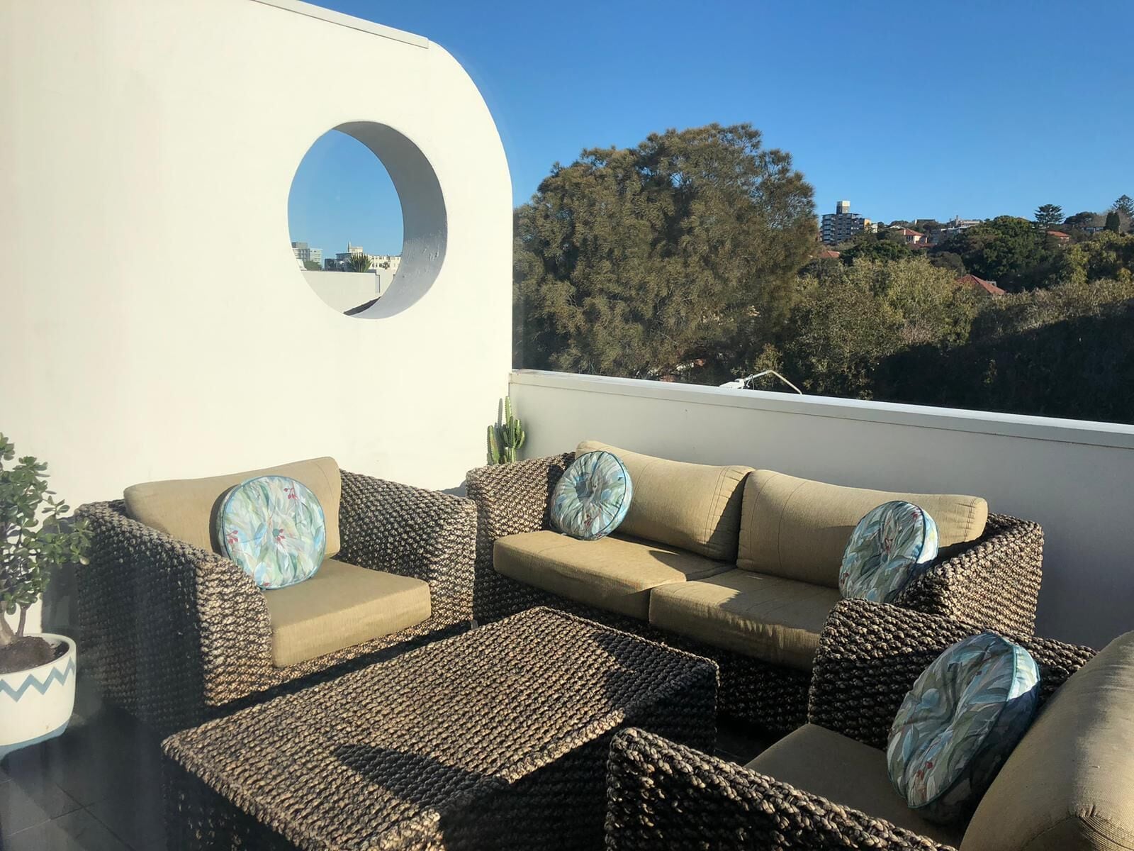 A rooftop terrace with wicker furniture, including a sofa and two armchairs, each with patterned cushions. A large circular window provides a view of the surrounding trees and buildings.