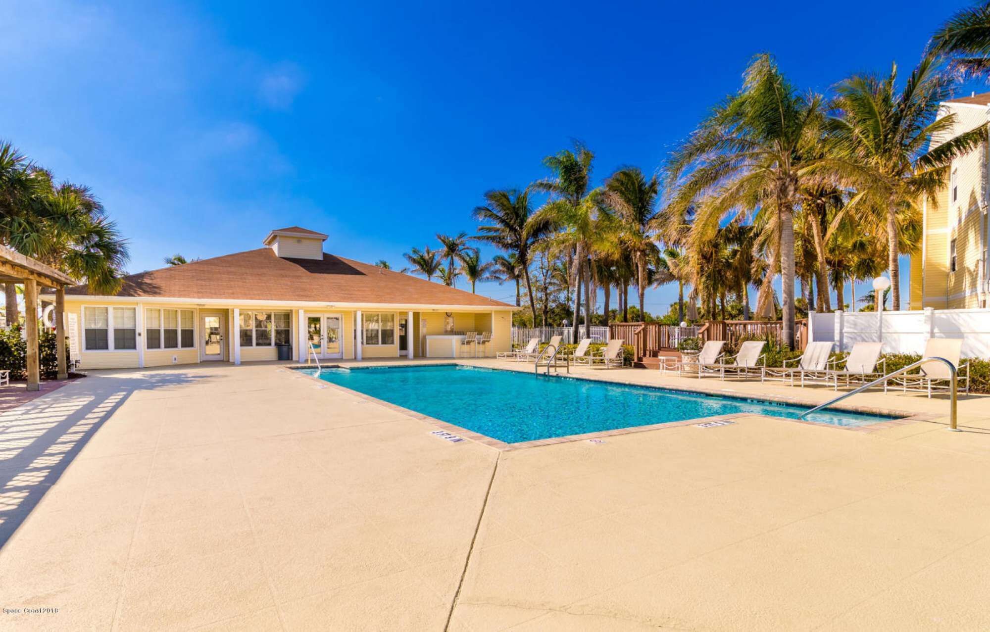 A pool area with lounge chairs, a building with a red roof, and palm trees.