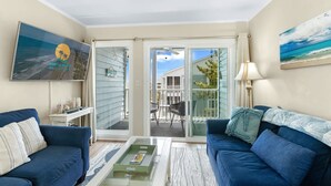 A living room with two blue sofas, a glass coffee table, a TV on the wall, and a sliding glass door leading to an outdoor patio. - Pelican Landing Unit 330 - Ocean view (Myrtle Beach)