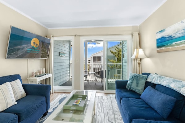 A living room with two blue sofas, a glass coffee table, a TV on the wall, and a sliding glass door leading to an outdoor patio.