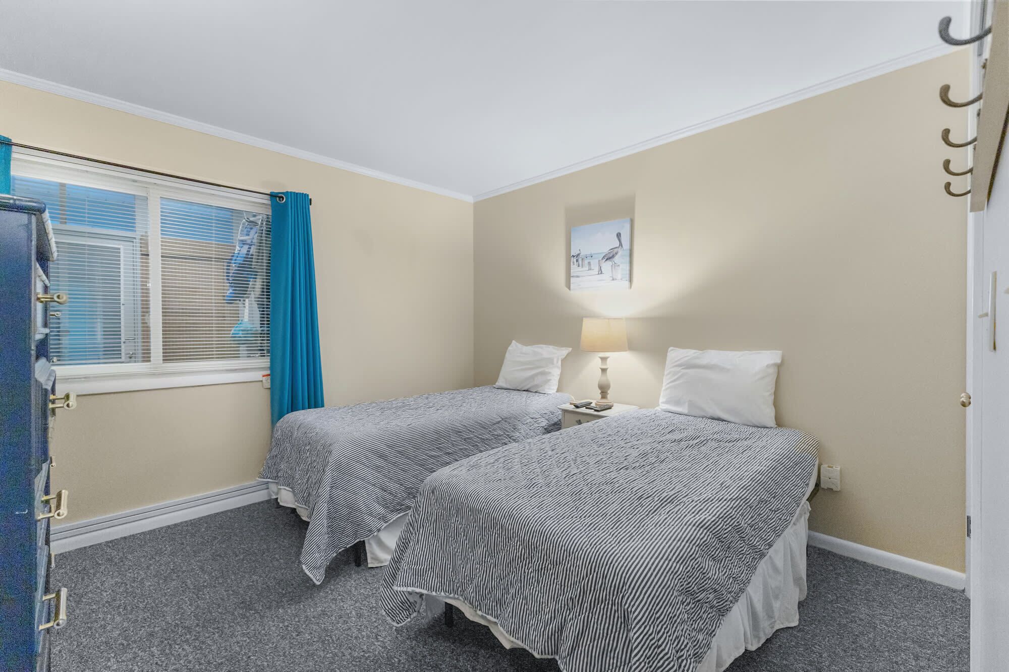 Une chambre avec deux lits simples, une fenêtre avec des rideaux bleus, une lampe fixée au mur et un tableau.