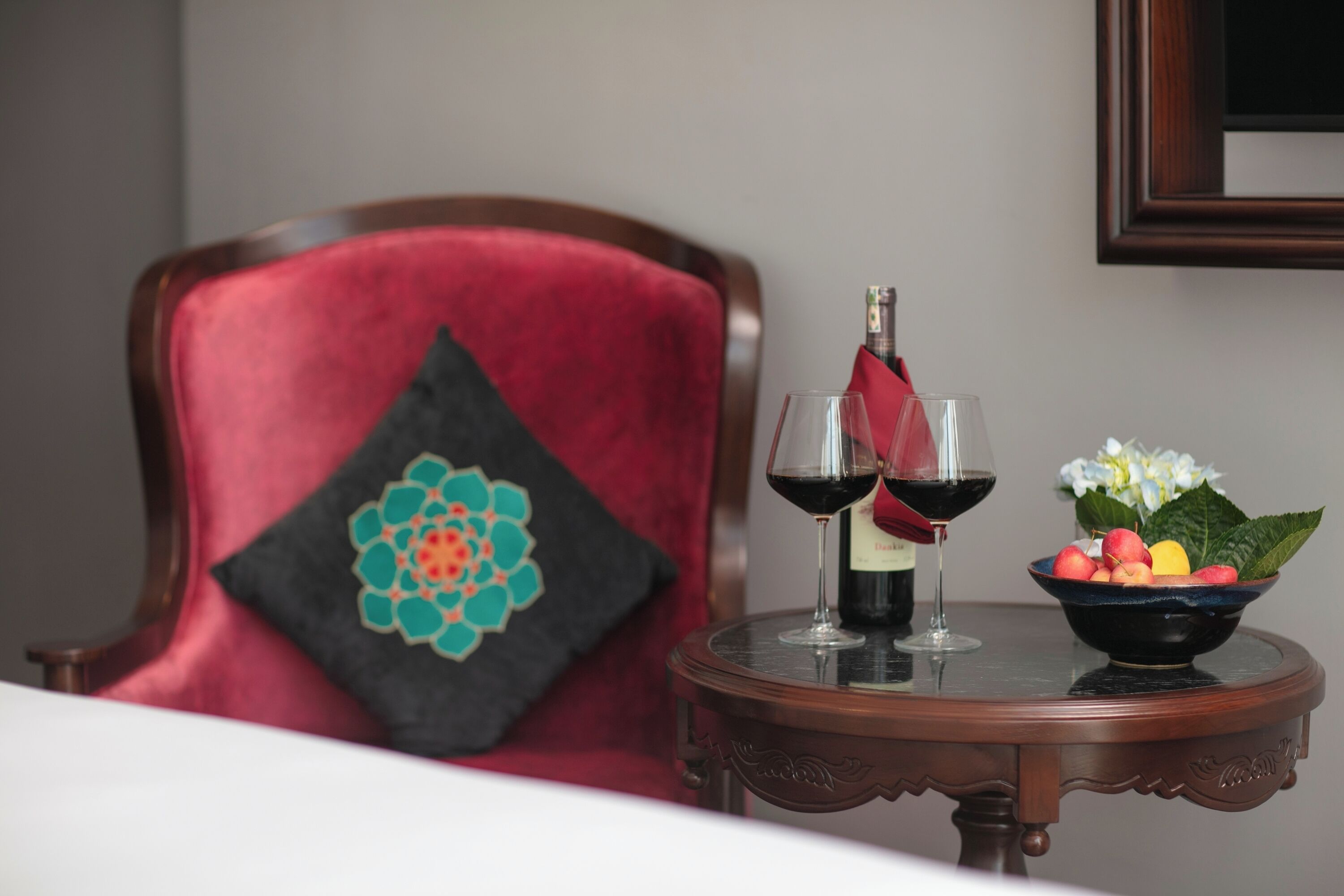 A room with a red chair, a small round table with a bottle of wine, two wine glasses, and a bowl of fruit.