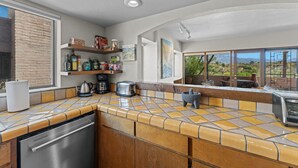 A kitchen with a dishwasher, coffee maker, toaster, and a stone mortar. There are shelves with food items and a window with a view of the outdoors.
