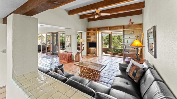 A spacious living area with a leather sofa, a coffee table, and a ceiling fan. There is a bookshelf and a TV in the background.