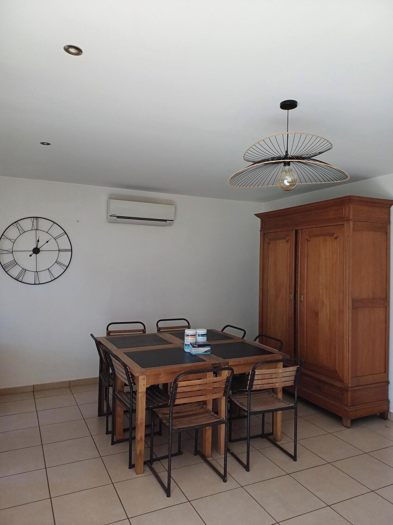 A dining area with a wooden table and chairs, a wall clock, an air conditioning unit, and a tall wooden cabinet.