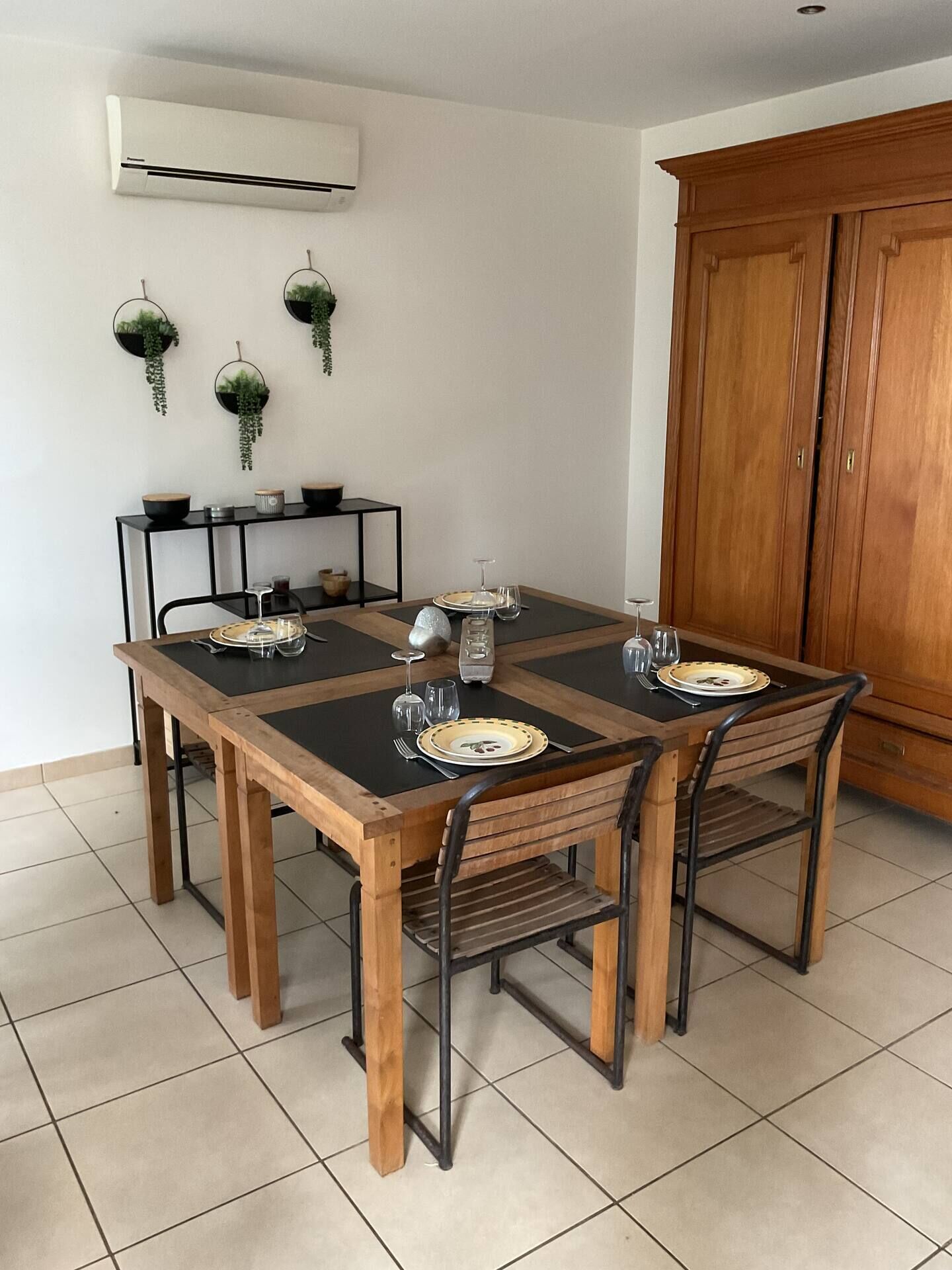 A dining area with a wooden table set for four, surrounded by chairs. There is a black metal shelf with decorative items and a white air conditioning unit on the wall.