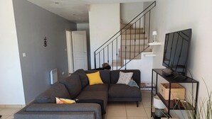 Ein Wohnzimmer mit grauem Ecksofa, einem Fernseher auf einem Standfuß, einer Treppe und einer Tür zu einem anderen Raum.