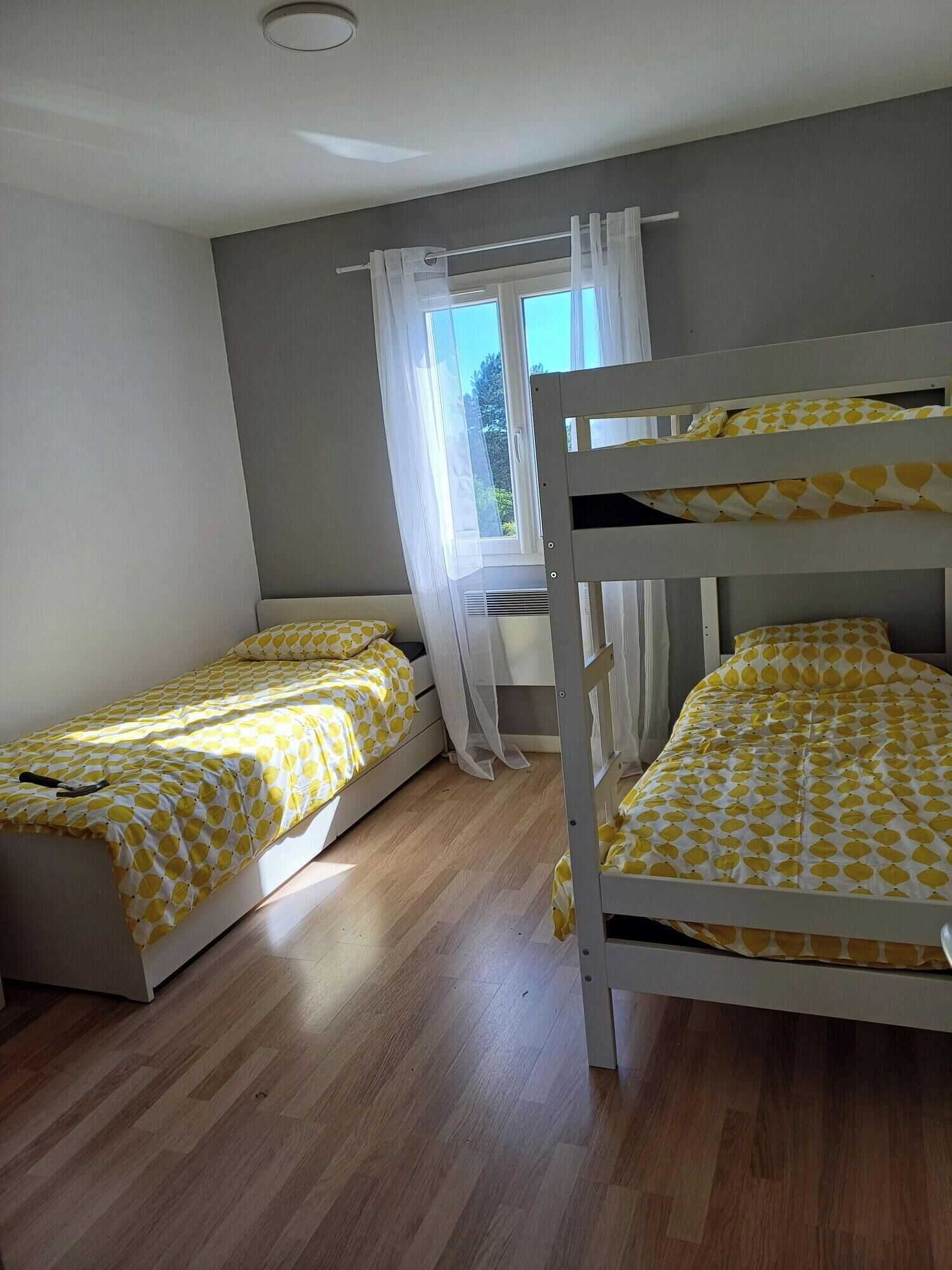A room with two bunk beds, each with a yellow and white patterned bedspread. There is a window with sheer curtains, and the room has wooden flooring.