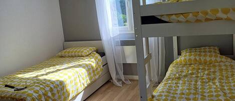 A room with two bunk beds, each with a yellow and white patterned bedspread. There is a window with sheer curtains, and the room has wooden flooring.