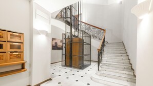 Una escalera con barandilla de metal y pasamanos de madera, que lleva a un rellano con pared blanca y un mueble de madera con puertas de vidrio.