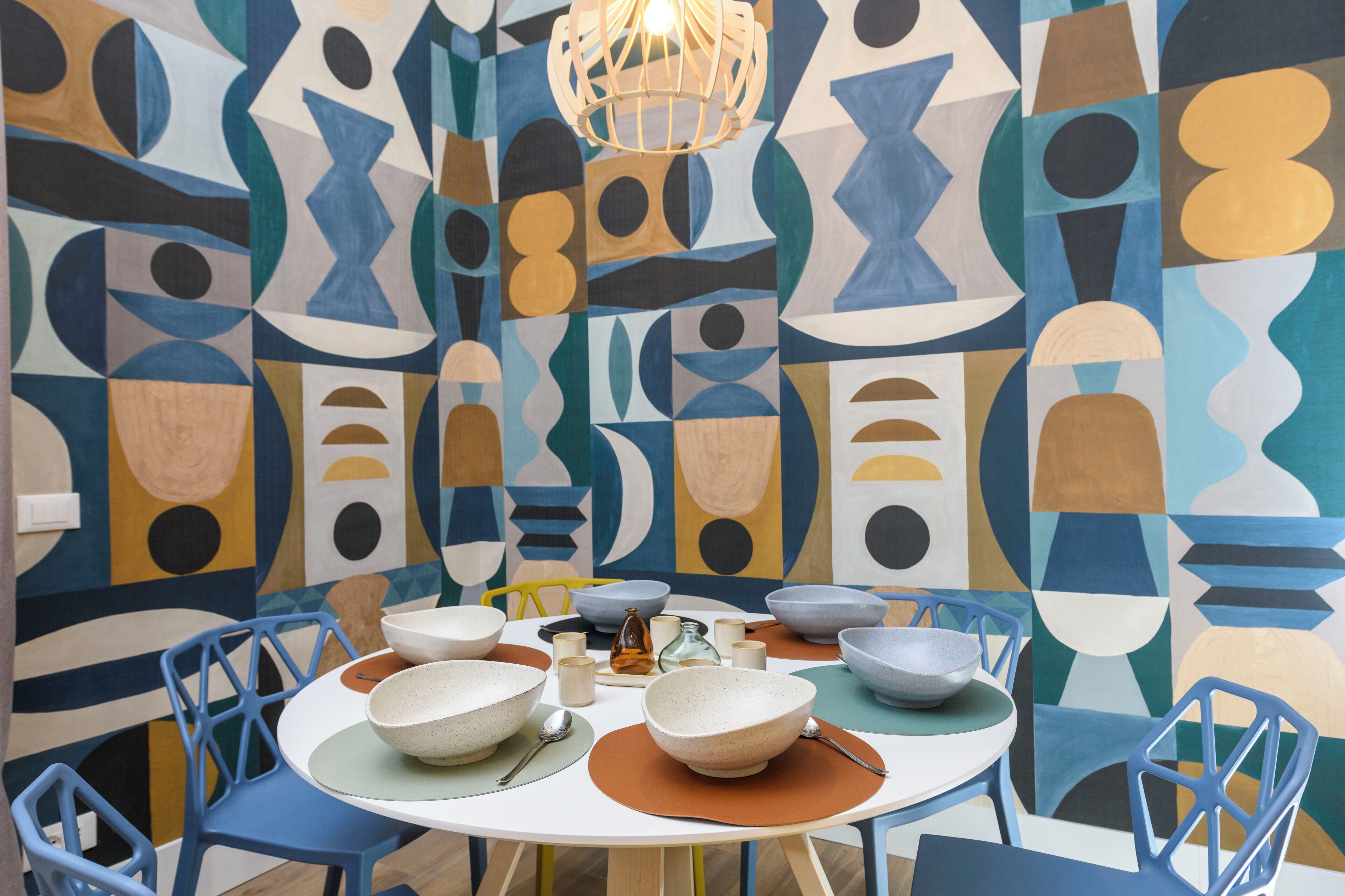 A dining area with a round table, blue chairs, and a geometric wall mural.