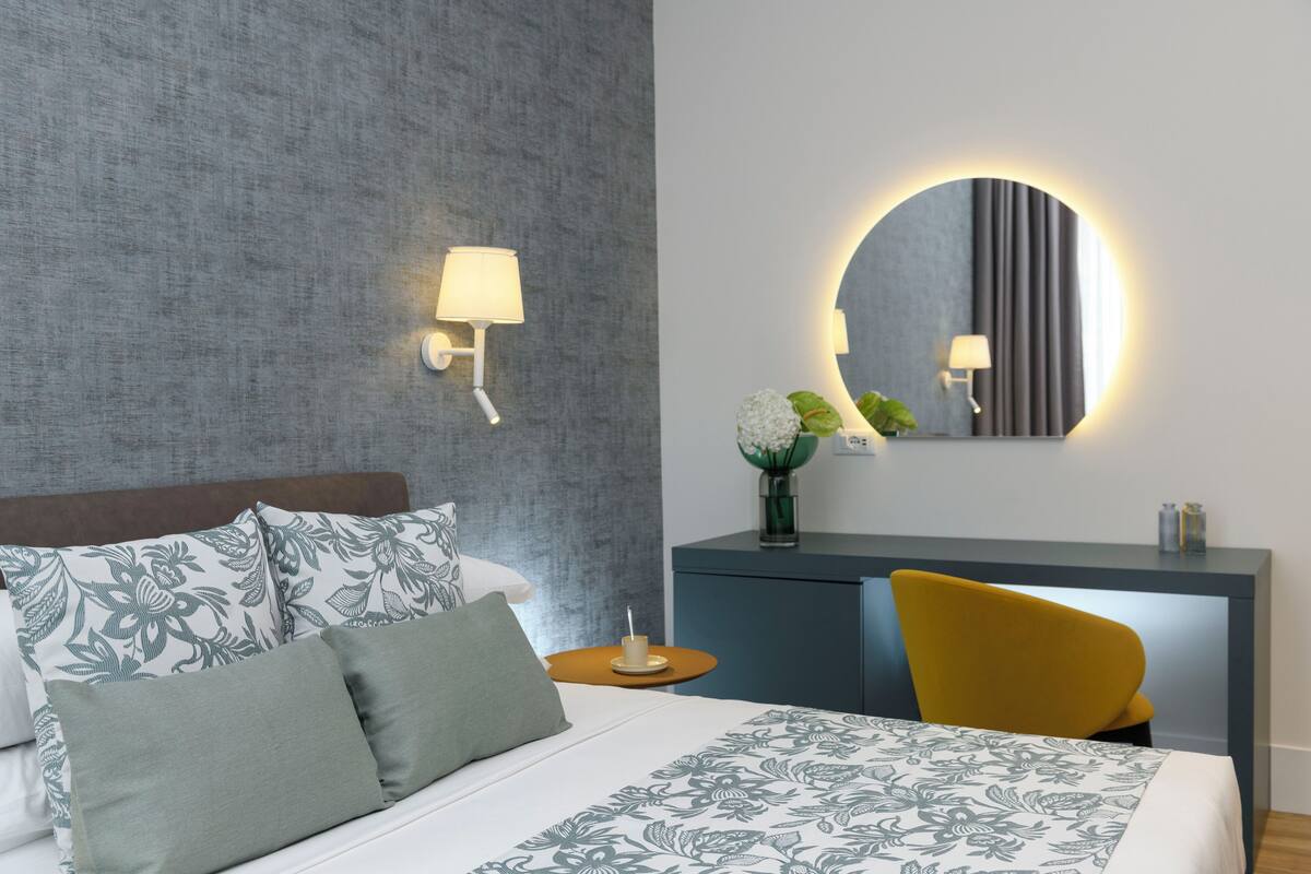 Ein ordentlich eingerichtetes Hotelzimmer mit einem Bett, einem Schreibtisch und einem Sessel. An den Wänden sind Lampen angebracht, und über dem Schreibtisch hängt ein Spiegel.