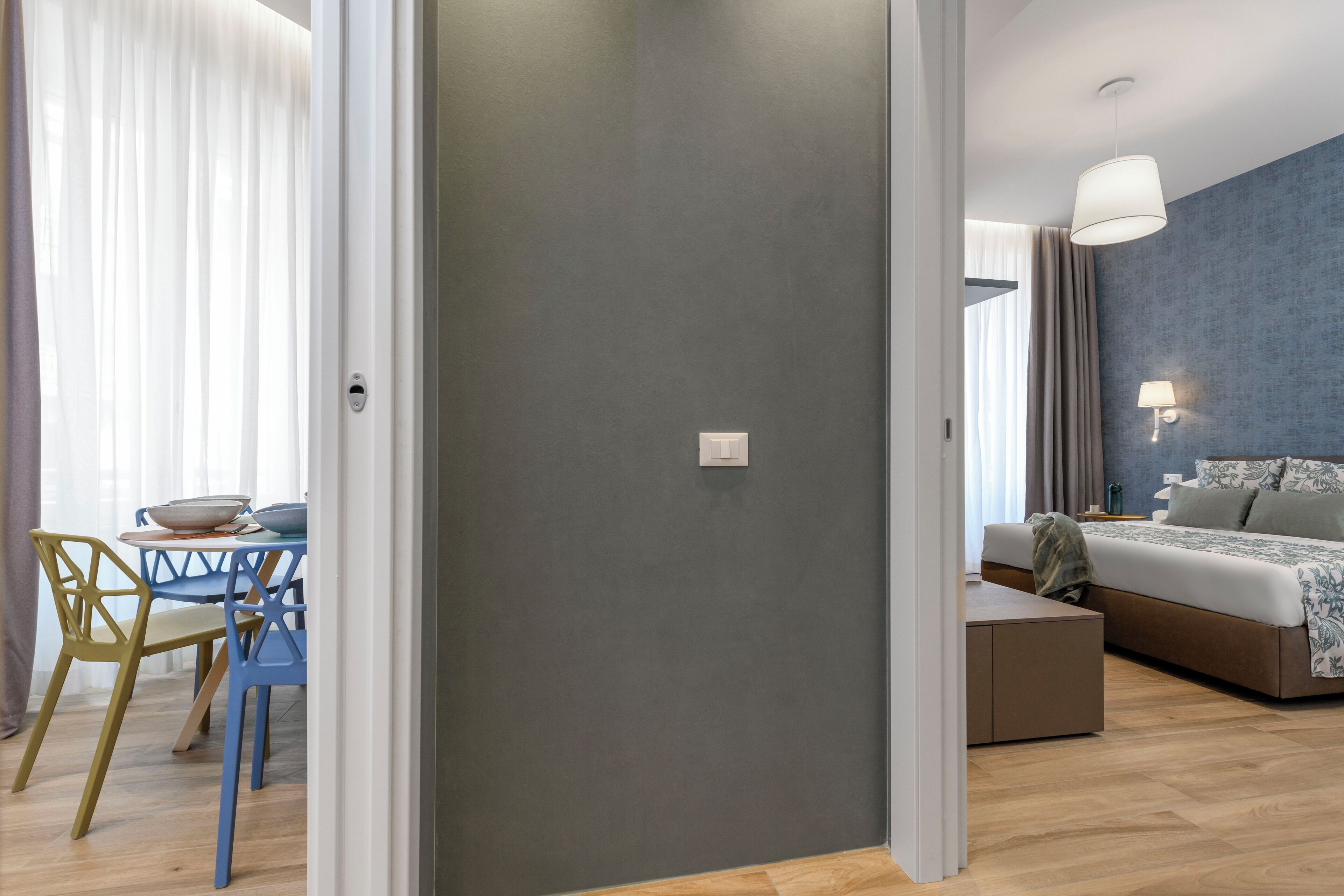 Een moderne hotelkamer met een grijze accentmuur, een bed met een beddegoed met een patroon, een nachtkastje en een lamp. Er is een eethoek met een tafel en stoelen zichtbaar door een open deur.