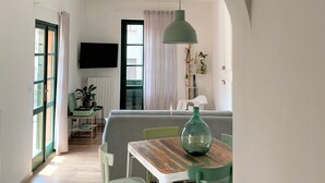 A dining area with a wooden table and chairs, a hanging light fixture, and a view through an archway to another room with a TV and plants.
