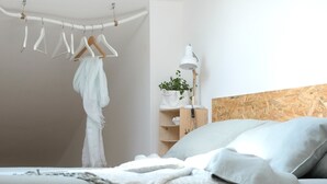 A bedroom with a loft bed, a hanging clothes rack, a bedside table with a lamp, and a potted plant.