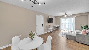 A living room with a dining area, a ceiling fan, and a TV mounted on the wall. - Stonecrest 620, 2Bd Apt I Free Parking I Pool (New Albany)