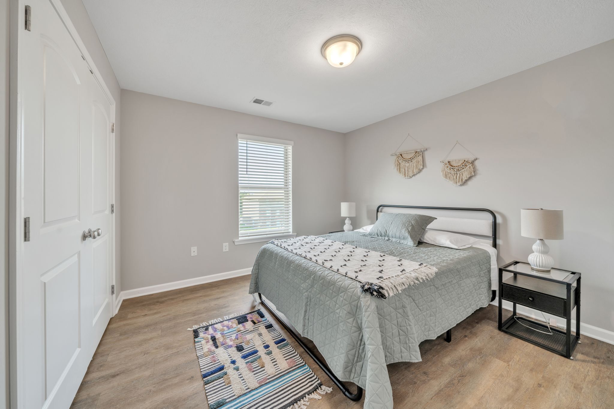 A bedroom with a bed, bedside tables, a window with blinds, and a door.