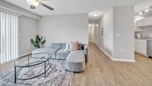 A modern living room with a grey sectional sofa, ottoman, and glass coffee table, a ceiling fan, and a kitchen in the background. - Stonecrest 712, 2Bd Apt I Free Parking I Pool (New Albany)