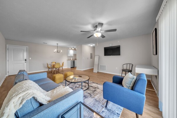 A modern living room with a blue sofa, a glass coffee table, and a ceiling fan.