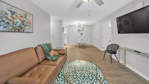 Un salon moderne avec un canapé en cuir marron, un tapis à motifs, un ventilateur de plafond, un coin repas avec une table et des chaises blanches, et un téléviseur fixé au mur.