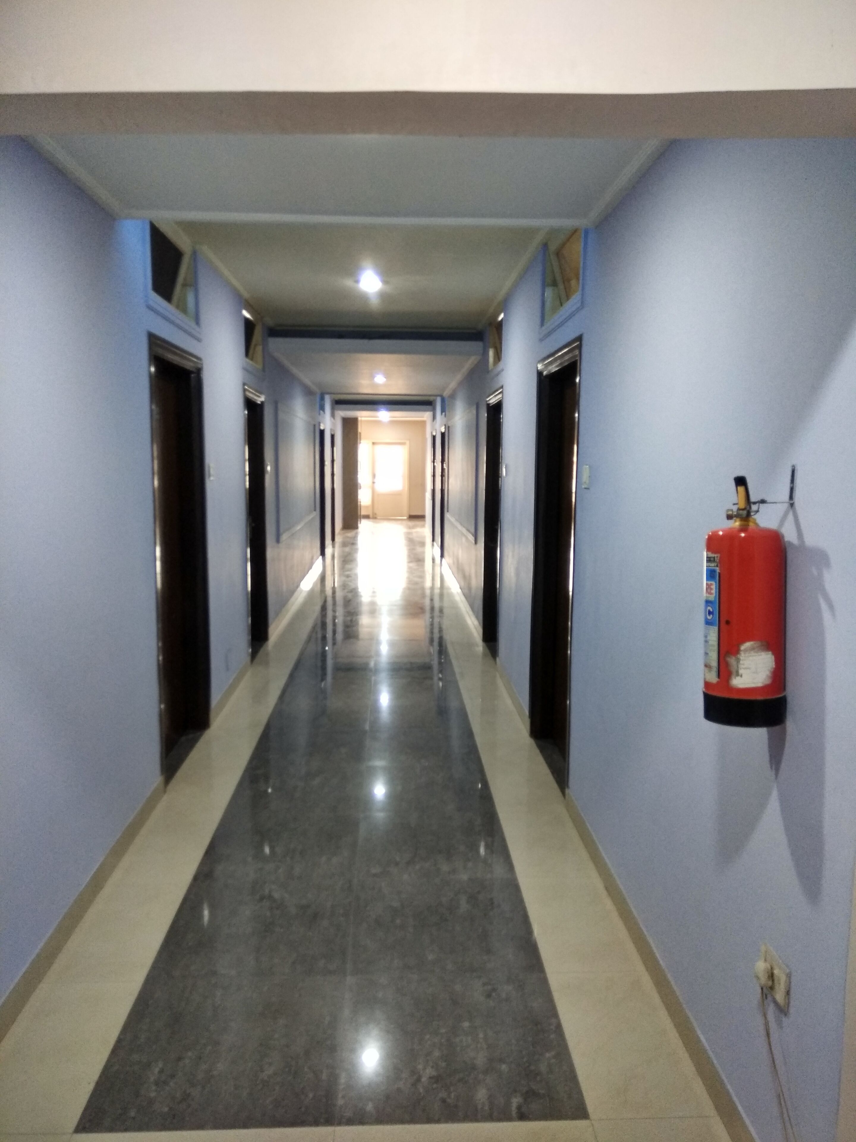 A long, narrow hallway with blue walls and a shiny floor, leading to a door at the end. A fire extinguisher is mounted on the wall to the right.