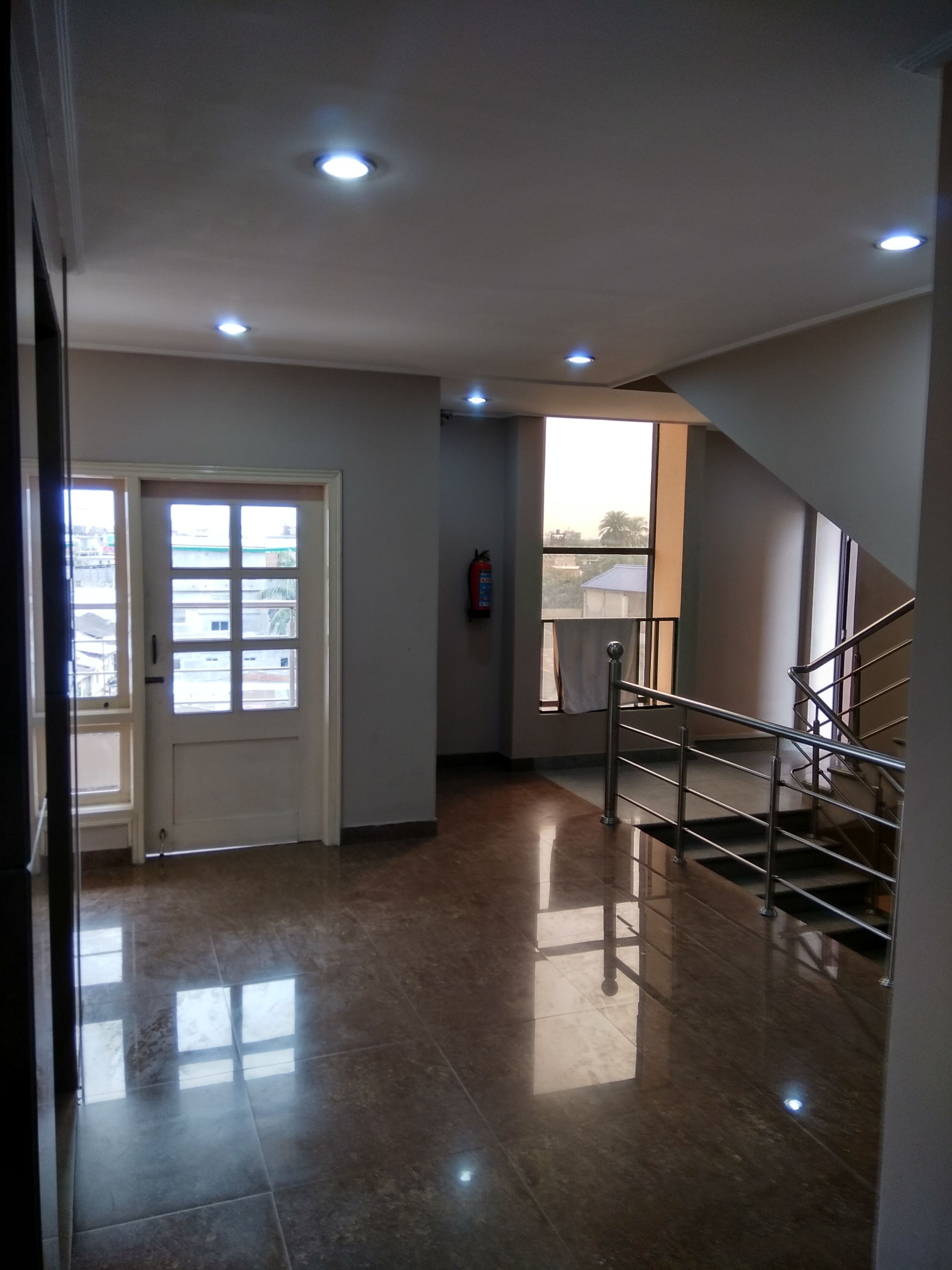 Un pasillo con una puerta de cristal, un extintor, una escalera y vistas de edificios exteriores.