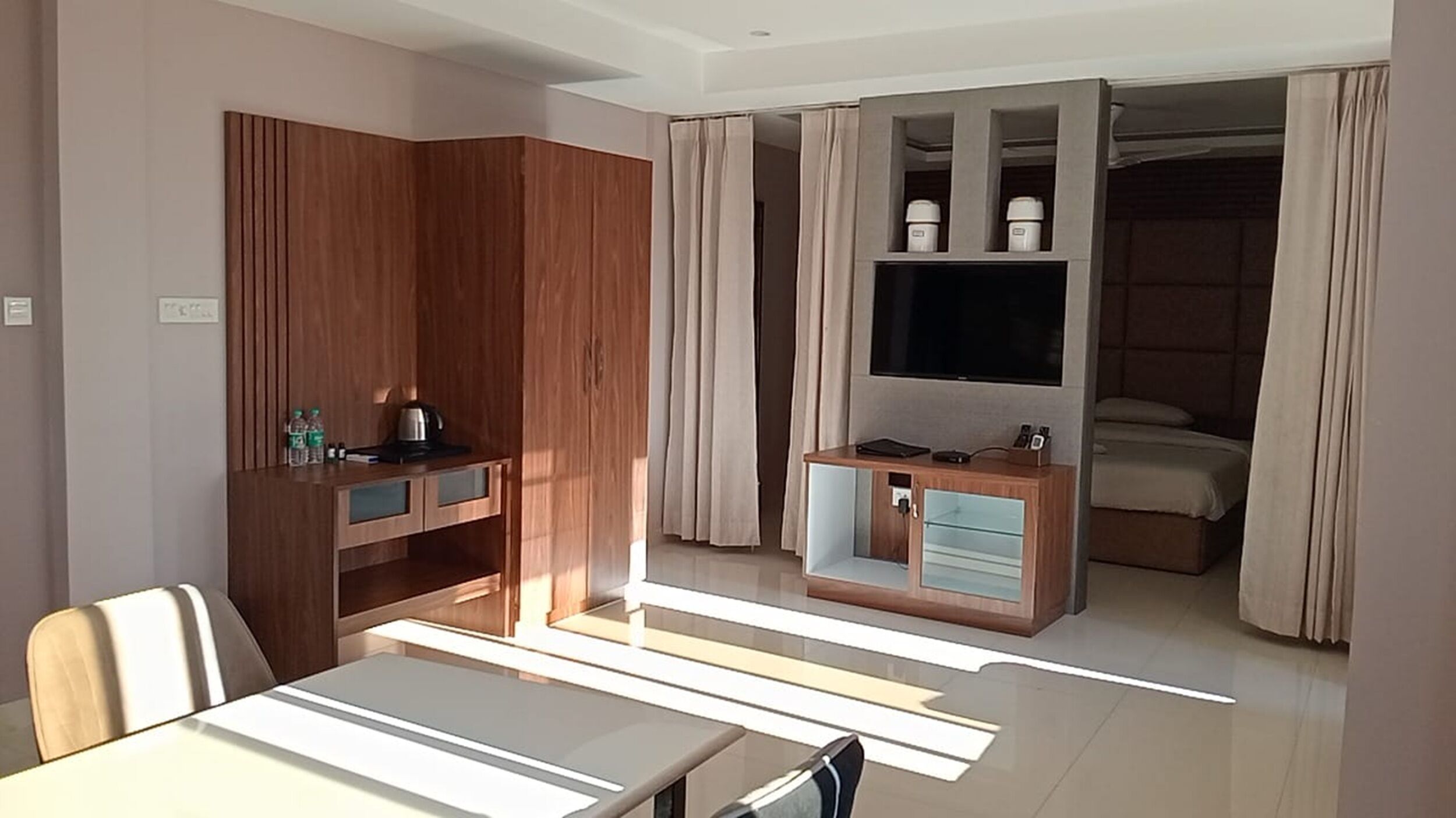 A modern hotel room with a wooden wardrobe, a dining table, a TV mounted on the wall, and a bed with curtains.