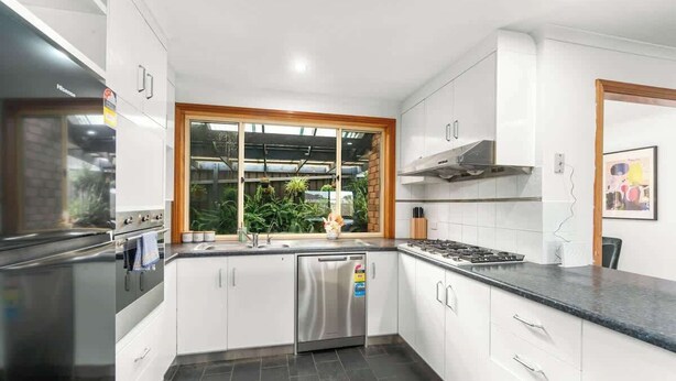 Cocina moderna con armarios blancos, lavavajillas de acero inoxidable, estufa a gas y ventana con vista a zonas verdes.
