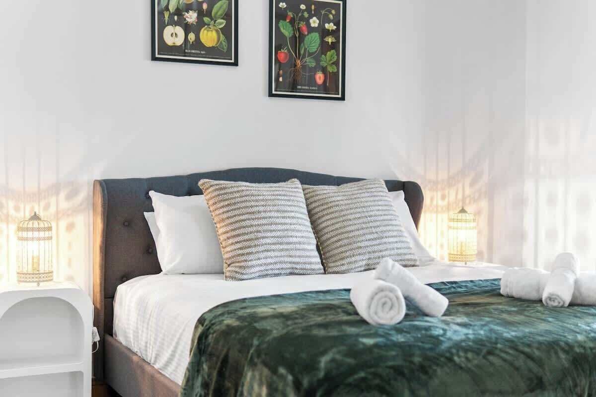 Uma cama bem arrumada com cabeceira escura, almofadas brancas e colcha verde. Há duas gravuras botânicas emolduradas na parede acima da cama.