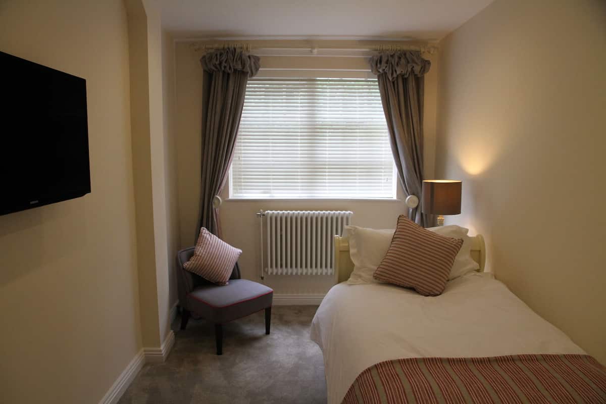 Une chambre d’hôtel avec un lit, une chaise, un téléviseur fixé au mur, une fenêtre avec des rideaux et un radiateur.