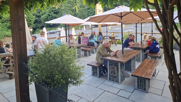 Une terrasse extérieure avec des tables en bois et des parasols, entourée de verdure et bordée par une route au loin.