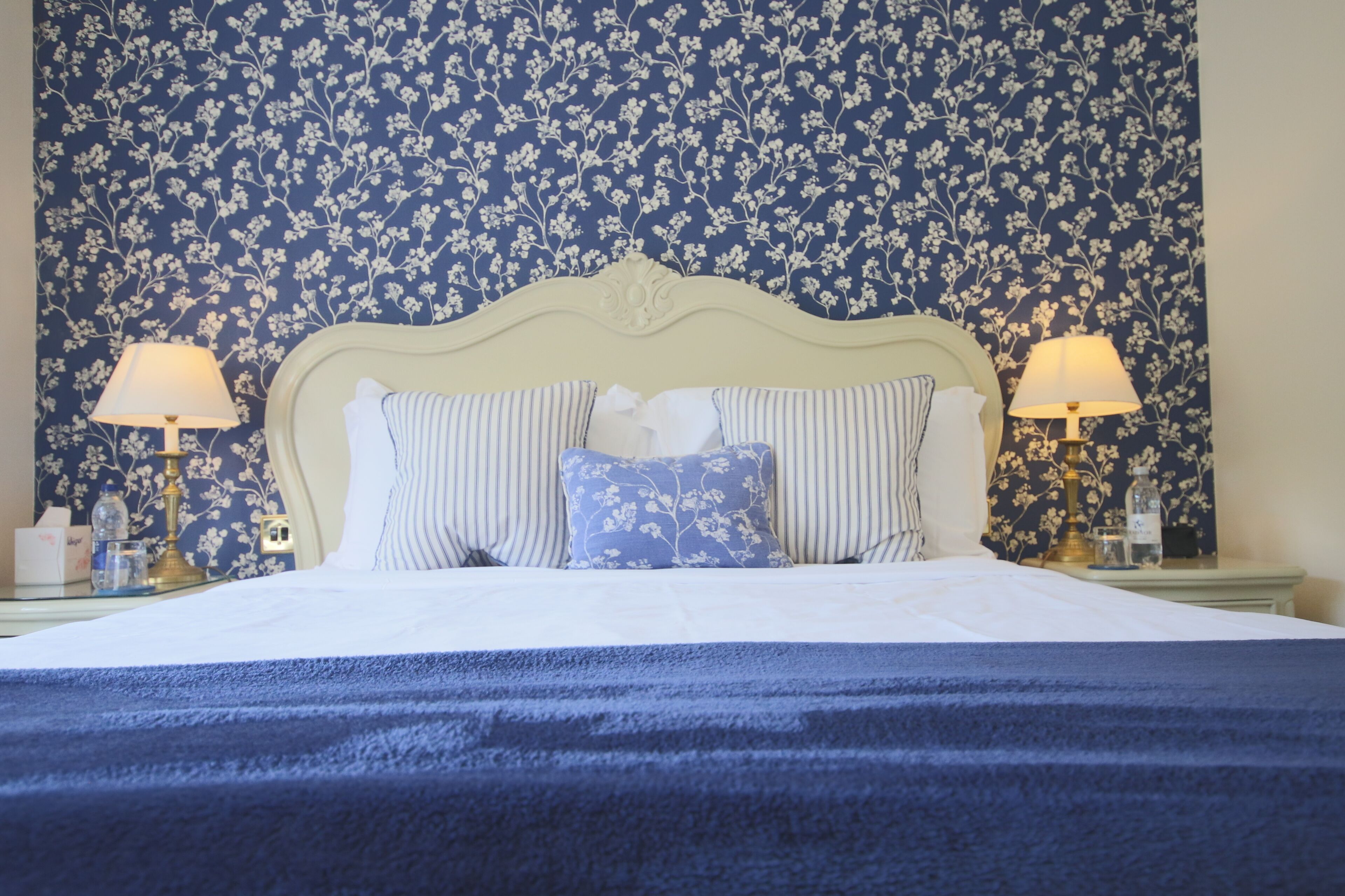 Een netjes opgemaakt bed met een blauwe deken, twee bedlampen en behang met bloemen.